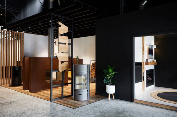 A dark, industrial-style open-plan interior featuring black exposed ceiling elements, timber slat screening, warm timber flooring, dark wood partitions, and a freestanding sculptural shelving divider. A compact lounge or media room is visible through the opening on the right, with a pale built-in unit and round rug. The main space includes a grey cylindrical fireplace or heater and a potted plant.