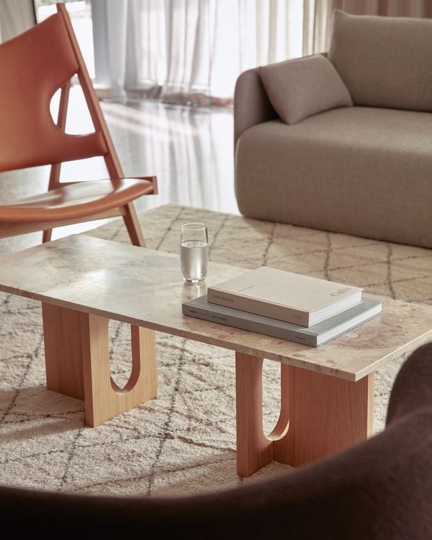 Warm, refined living room vignette featuring a low rectangular coffee table with a pale veined stone top and sculptural light-wood U-shaped legs. A tan upholstered sofa, matching lounge chair, patterned neutral rug, sheer curtains, stacked books, and a glass of water create a calm contemporary setting.