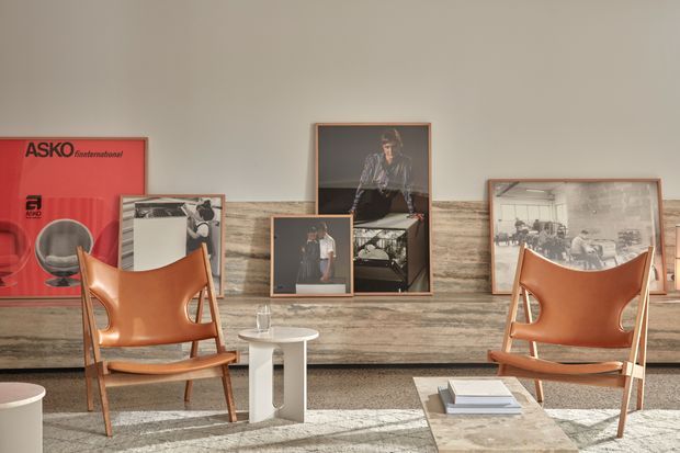 A warmly styled mid-century-inspired living room with two sculptural brown leather lounge chairs, a small white side table, and a low stone or concrete coffee table. Framed photographic artwork and a large red poster are arranged against a horizontally textured timber or stone feature wall. Neutral walls, earthy materials, and strong sunlight create a refined, curated atmosphere.