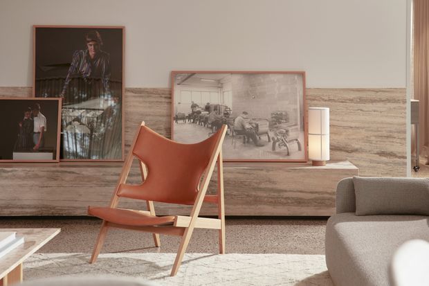 A warmly lit contemporary living room vignette featuring a cognac leather sling lounge chair with a light timber frame, a pale upholstered sofa, and a low marble or stone coffee table. A full-width horizontally veined stone feature wall and matching ledge form the backdrop, displaying framed sepia artwork and photographs alongside a slim cylindrical lamp. Neutral textiles, timber accents, and soft daylight create a refined, gallery-like atmosphere.