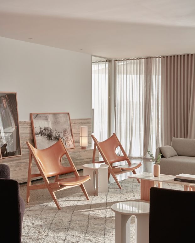 A softly lit contemporary living room with floor-to-ceiling sheer curtains, a neutral sofa, sculptural white side tables, a pale patterned rug, and two warm tan leather sling lounge chairs. Framed artwork leans against a textured neutral wall, while a low timber coffee table and small plants add warmth. The room has a calm, refined, light-filled atmosphere.