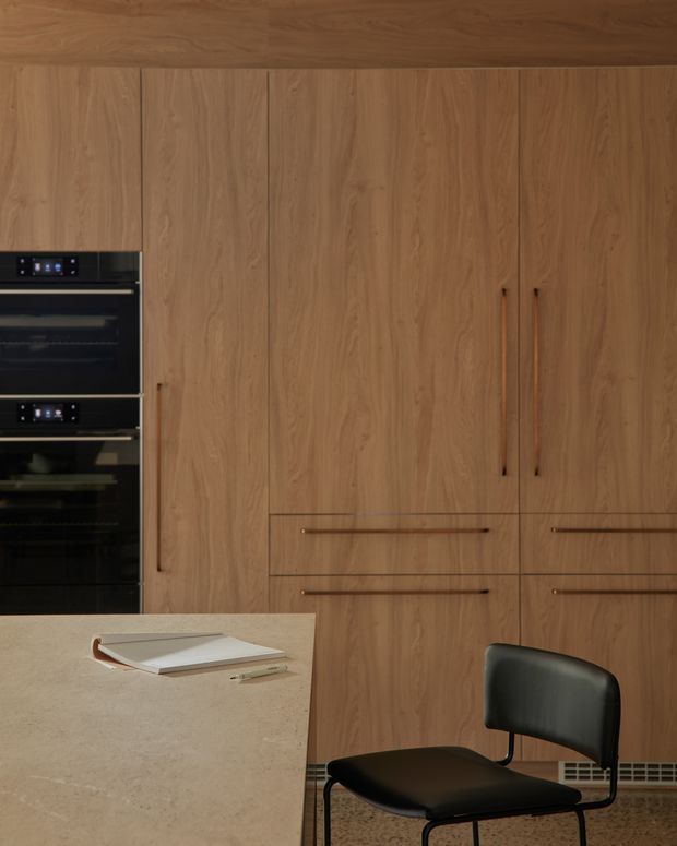 A close-up view of a refined contemporary kitchen featuring full-height warm timber cabinetry with integrated drawers and tall storage, stacked black built-in ovens, and a pale stone island or benchtop in the foreground. A simple black dining chair and a notebook add scale, while the restrained material palette creates a warm, sophisticated atmosphere.