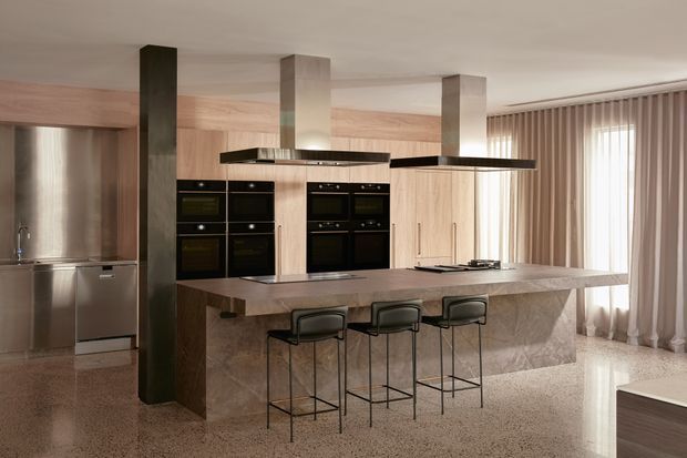 A spacious contemporary kitchen with a large stone island and three slim bar stools. The room features extensive light timber cabinetry, integrated black ovens and appliances, dark structural columns, twin suspended rangehoods, floor-to-ceiling curtains, and a polished terrazzo floor. Warm neutral finishes and abundant filtered daylight create a refined, modern atmosphere.
