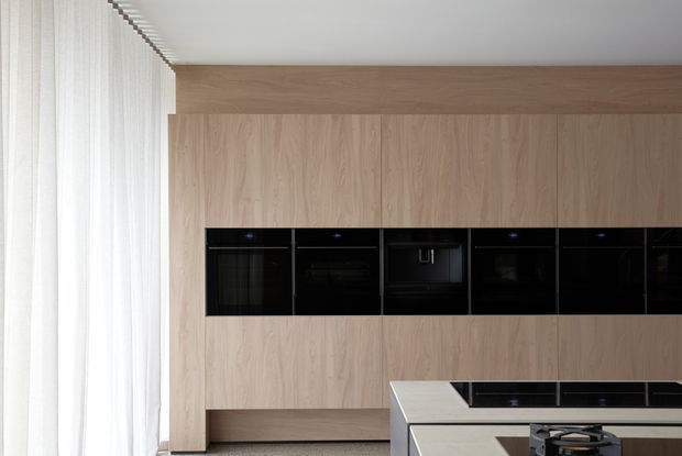 Contemporary minimalist kitchen with a full-height light timber wall and integrated black appliance bank. A pale stone island with a black cooktop occupies the foreground, while sheer white curtains provide soft natural light along the left side.