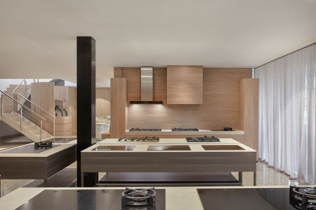 A spacious contemporary kitchen with multiple cooking islands and a long rear wall of warm timber cabinetry. The foreground island features dark cabinetry, pale stone worktops, integrated gas burners and cooktops, while a black structural column and open staircase with metal balustrade are visible to the left. Full-height sheer curtains line the right side, creating a bright, refined interior.