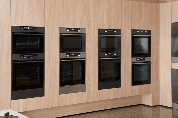A contemporary kitchen appliance wall featuring multiple built-in black ovens and compact ovens set into full-height light timber cabinetry. The cabinetry forms a clean, symmetrical composition, with a visible terrazzo-like floor and a partial glimpse of additional cabinetry at the right edge.