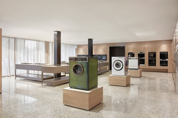 A bright, spacious modern appliance showroom with a polished terrazzo floor, timber-look wall cabinetry, and large sheer-curtained windows. Washing machines, ovens, and other appliances are displayed on light timber plinths and integrated into fitted kitchen-style joinery. A central island and dark structural columns define the open-plan commercial space.