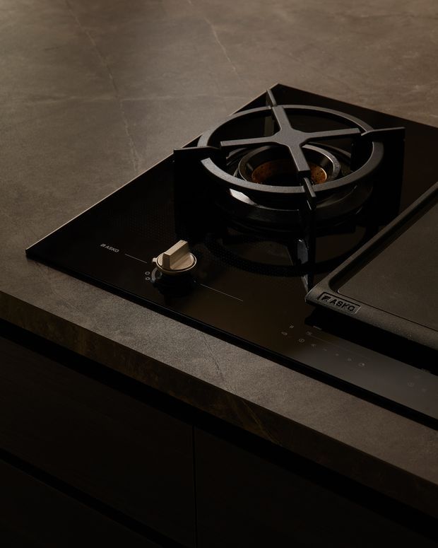 Close-up product-style image of a black built-in gas cooktop set into a dark stone kitchen benchtop. The cooktop has a single visible heavy-duty burner with cast-iron trivets, a metallic control knob, and glossy black glass surfaces. Dark cabinetry is visible below, creating a moody contemporary kitchen setting.
