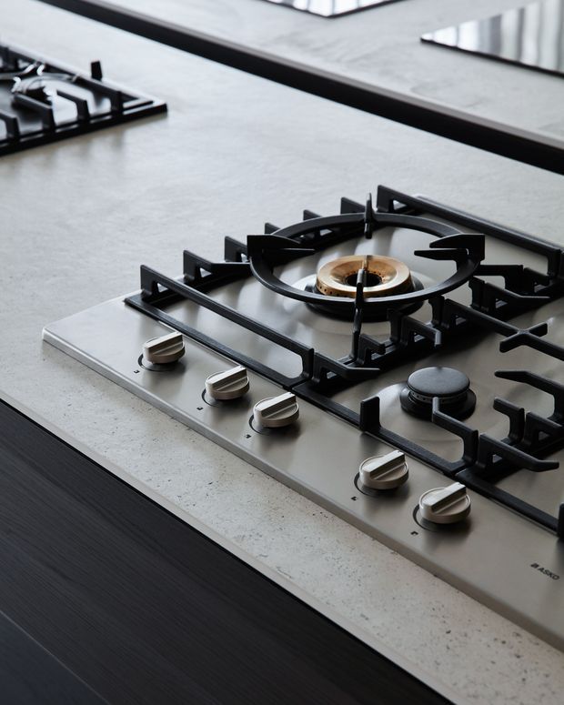 Close-up product-style photograph of a built-in stainless-steel gas cooktop set into a pale stone or engineered-stone kitchen benchtop. Multiple black cast-iron pan supports, brass-toned burners, and metallic control knobs are visible, with dark cabinetry below and a reflective splashback or rear surface in the background.