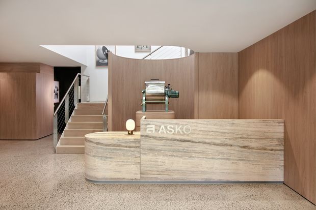 A contemporary commercial reception area with a curved stone reception desk bearing ASKO branding, a polished industrial machine displayed behind it, warm timber wall panelling, terrazzo flooring, and a stairway leading to an upper level. The space is softly lit with a clean, refined material palette.