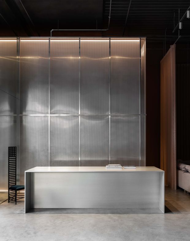 A moody contemporary interior featuring a large translucent corrugated-panel partition framed in metal, a minimalist pale reception or service counter, polished concrete flooring, a black exposed ceiling, and warm linear lighting. A black chair and a partially visible lounge area appear at the sides.