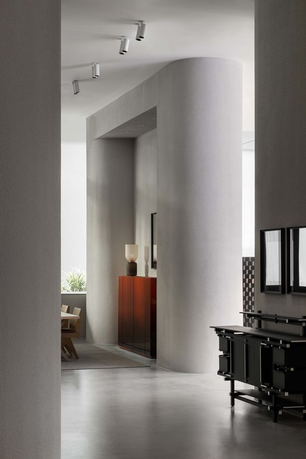 A refined contemporary interior viewed through a doorway, featuring sculptural curved plaster walls, a smooth grey floor, recessed and track lighting, and a striking black built-in kitchen or bar joinery unit in the foreground. A red-orange sideboard and dining area are visible beyond the curved partition. The space has a restrained monochromatic palette with strong geometric and curved forms.