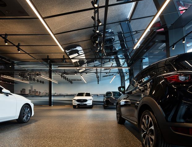 Wide-angle interior photograph of a modern automotive showroom or dealership, with several cars displayed on a polished speckled floor. The space features a dark exposed ceiling grid, extensive reflective glazing or mirrored panels, black structural framing, and linear LED strip lighting, creating a sleek, high-contrast commercial environment.