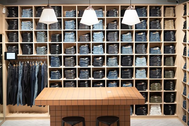 A well-organised clothing retail space with floor-to-ceiling open timber shelving filled with neatly folded jeans and garments. Hanging denim is displayed along the left side, while a tiled central display counter with two stools occupies the foreground. Three large pendant lights illuminate the warm, minimalist shop interior.