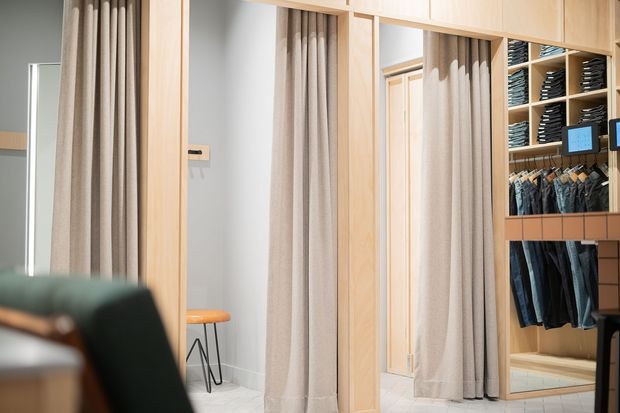 A contemporary clothing retail or wardrobe display area with light timber-framed fitting-room partitions, full-length beige curtains, pale tiled flooring, and open shelving displaying folded garments and hanging jeans. A small stool and partial seating are visible in the foreground.