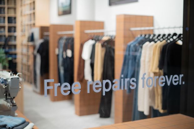 A softly focused clothing retail or tailor shop interior viewed through a glass storefront, with multiple racks of hanging garments, warm wood display panels, shelving, and a sewing machine in the foreground. Large lettering on the glazing reads “Free repairs forever.”