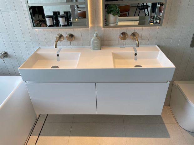 Contemporary bathroom vanity with a wide white double basin, wall-mounted white cabinetry, brushed metal wall-mounted taps, large mirrored cabinets, and light grey tiled walls and floor. A bathtub is partially visible on the left and a toilet on the right.