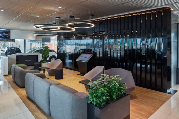 A contemporary automotive showroom or dealership reception lounge with a curved grey sectional sofa, mauve lounge chairs, round side tables, indoor plants, and a central display kiosk. The space features warm timber flooring, glossy black vertical wall fins, extensive glazing, a slatted acoustic ceiling, and large circular illuminated pendant lights. Vehicles are visible in the showroom beyond the lounge.