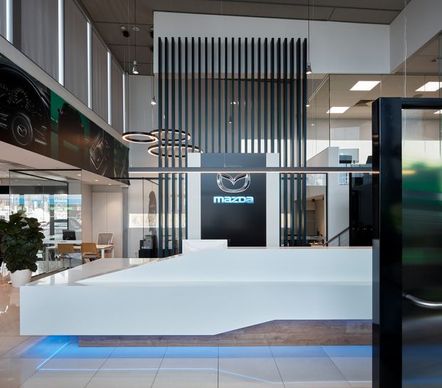 A contemporary Mazda automotive showroom and reception area with a large white sculptural reception desk illuminated by blue underlighting. The space features a double-height central volume with vertical dark timber or metal slats, glazed partitions, pendant ring lights, polished light tile flooring, and a prominent Mazda logo wall. Vehicle imagery and showroom displays are visible along the sides.