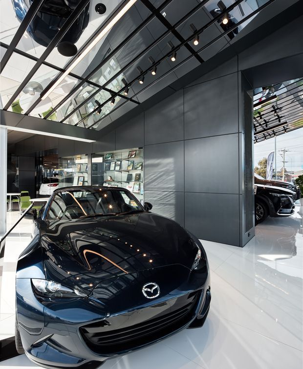 A contemporary automotive showroom interior featuring a glossy black Mazda sports car prominently displayed on a polished white floor. The space has dark charcoal wall panels, extensive glass and mirrored surfaces, exposed black ceiling framing, track lighting, linear illuminated lighting, and large glazed openings with additional vehicles visible outside. Bright daylight and reflections create a high-contrast, polished commercial environment.