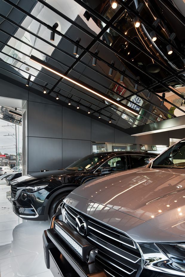 Interior view of a contemporary luxury car dealership showroom, with several vehicles displayed beneath a dramatic glossy black exposed ceiling grid, extensive reflective ceiling panels, track lighting, linear LED lighting, and dark wall cladding. Large front glazing provides street visibility, while the polished light floor and reflective surfaces create a high-contrast commercial environment.