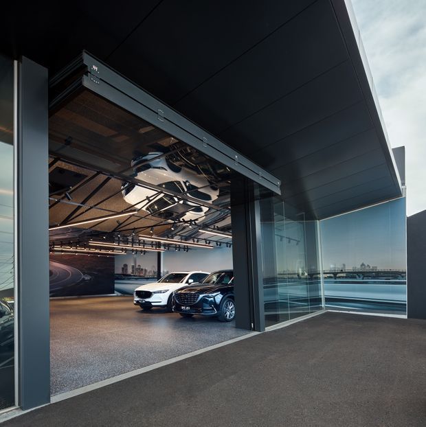 Exterior view into a contemporary automotive showroom or commercial display space through a large open sectional glass-and-metal doorway. Several cars are displayed inside beneath a dramatic dark ceiling with exposed suspended structural elements and track lighting. Full-height glazing on the right reflects the surrounding urban setting, while the broad projecting roof canopy and dark charcoal finishes create a sleek, industrial-modern appearance.