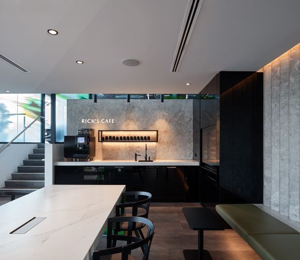 A contemporary commercial cafe interior with a large pale stone island and bar seating in the foreground. The rear service counter features dark cabinetry, a pale stone backsplash, integrated coffee equipment, pendant-free illuminated shelving, and signage reading “RICK’S CAFE.” Black cabinetry continues along the right wall beside a green upholstered banquette and small tables. Concrete-textured wall panels, polished timber-toned flooring, exposed stair access, and extensive glazing create a refined urban hospitality setting.