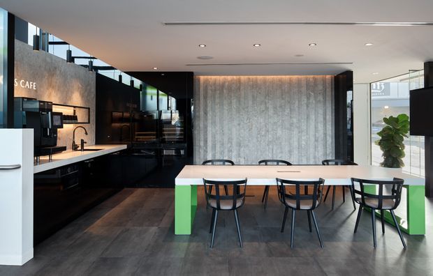 Contemporary open-plan café or commercial dining space with a long communal table, black chairs, dark kitchen or service joinery, integrated coffee equipment, concrete-look feature wall, and large glazed openings. Bright green table supports and indoor planting add colour to the otherwise monochrome interior.