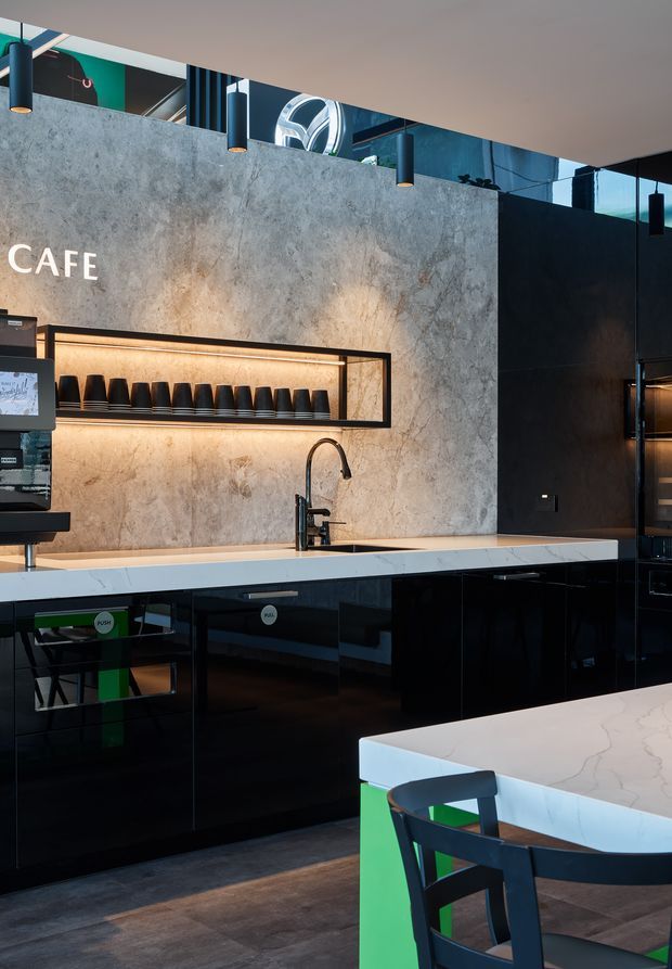 A contemporary café interior with a compact service kitchen and counter. Glossy black cabinetry runs beneath pale marble-look benchtops, while a textured grey feature wall incorporates an illuminated display shelf with coffee cups and the word “CAFE.” Black pendant lights, dark flooring, green accents, and café seating create a polished, high-contrast commercial fit-out.
