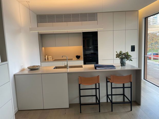 Contemporary minimalist kitchen with a large pale island, integrated cabinetry, concealed appliances, warm under-cabinet lighting, timber flooring, and two black-framed stools with tan leather seats. Full-height glazing opens toward an outdoor deck, while a linear ceiling detail adds subtle architectural interest.