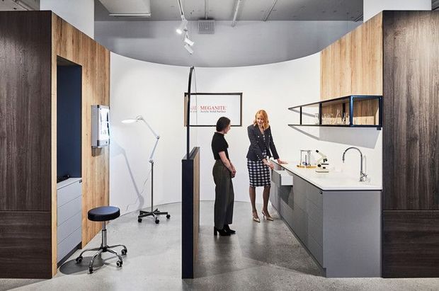Contemporary kitchen showroom or design studio with a compact display kitchen, white curved feature wall, dark wood and timber-look cabinetry, open shelving, integrated appliances, a polished concrete floor, and track lighting. Two people are present, with a small consultation or work area visible to the left.