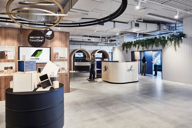 A contemporary commercial showroom or reception space with a curved black reception counter, product display plinths, wood-panelled branding wall, pale concrete-look flooring, and a sculptural white service desk. Exposed ceiling services, track lighting, suspended circular feature lights, and hanging greenery contribute to an industrial yet polished fit-out.