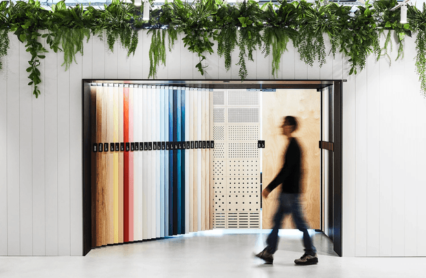 A bright contemporary commercial showroom or architectural materials display space. A large pivoting opening in a white vertically panelled wall reveals tall samples of timber, coloured panels, and perforated metal finishes. A blurred person walks across the smooth light-coloured floor, while abundant hanging greenery forms a lush overhead border.