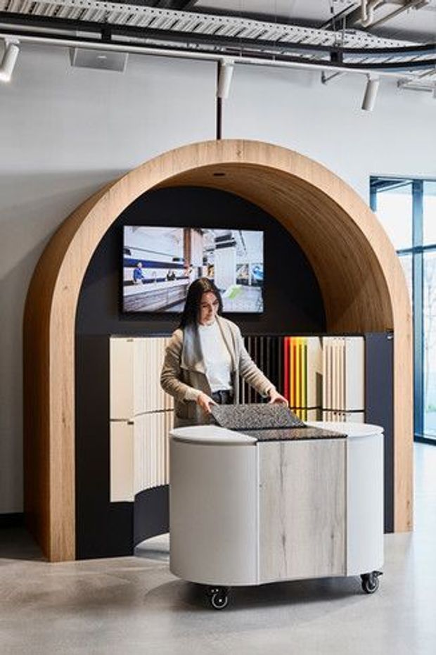 A contemporary commercial showroom or design studio interior featuring a large rounded timber arch framing a black display wall with a mounted screen and vertical material samples. In the foreground is a mobile curved display or work counter with casters, while exposed services, track lighting, and glazing reinforce the open industrial-commercial setting.