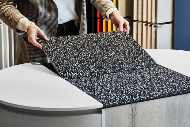 A close-up product-style image showing a person presenting a large black-and-white speckled surface sample over a curved white counter and adjacent pale wood-grain cabinetry. Vertical material samples are visible in the background, suggesting a kitchen or interior finishes showroom.