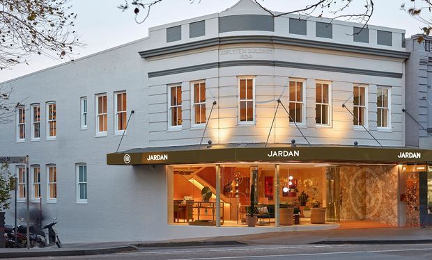 Street-facing view of a renovated multi-storey commercial building with a pale painted masonry facade, symmetrical tall windows, a projecting cornice, and a small curved-roof central upper section. The ground floor contains a warmly lit retail storefront with dark branded awnings and large display windows.
