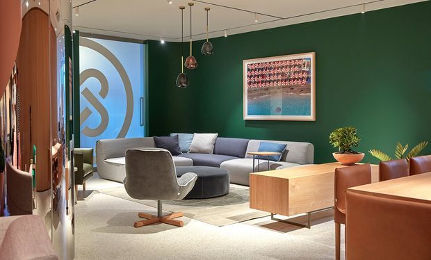 A refined contemporary living room with a large curved sectional sofa, sculptural swivel chair, round coffee ottoman, timber storage cabinet, and leather dining chairs. Deep green walls contrast with light neutral carpet and upholstery, while pendant lights and framed artwork add visual interest. A curved glazed or decorative partition is visible at the left.