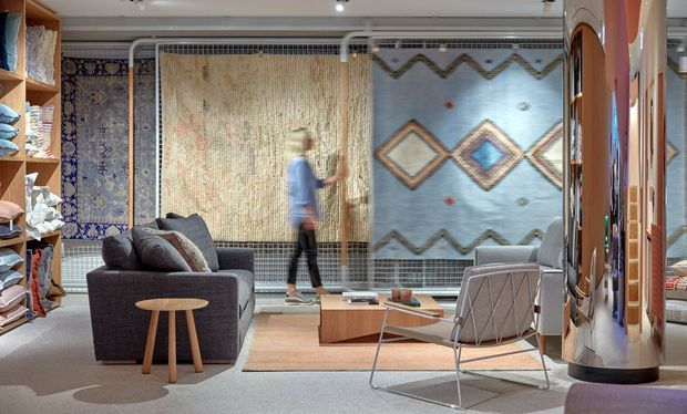 A contemporary textile showroom or display space with layered rugs and woven wall hangings suspended behind mesh frames. The room includes a dark grey sofa, a low timber coffee table, a sculptural lounge chair, open shelving with folded textiles, polished reflective display columns, and recessed ceiling lighting. The overall palette is neutral and earthy with geometric textile patterns creating visual accents.