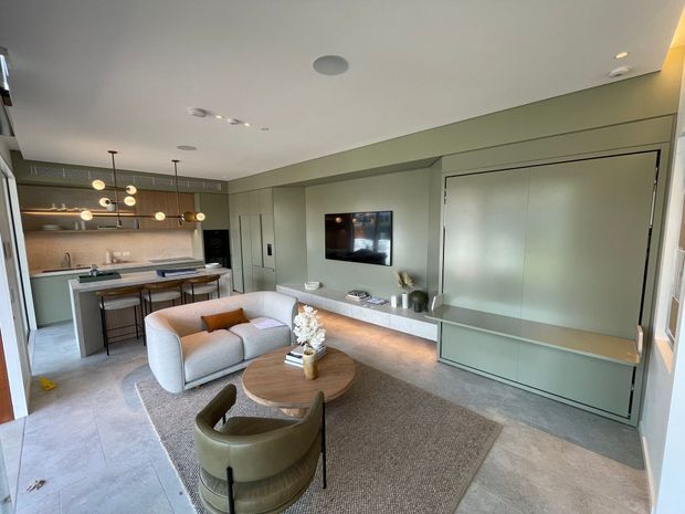 Contemporary open-plan living room with an integrated kitchen at the rear. The space features pale grey-green built-in cabinetry and wall panelling, a wall-mounted television, a long illuminated media console, a compact pale sofa, curved lounge chair, round timber coffee table, neutral rug, and understated pendant and recessed lighting. Large-format pale flooring and a restrained neutral palette create a refined, cohesive interior.