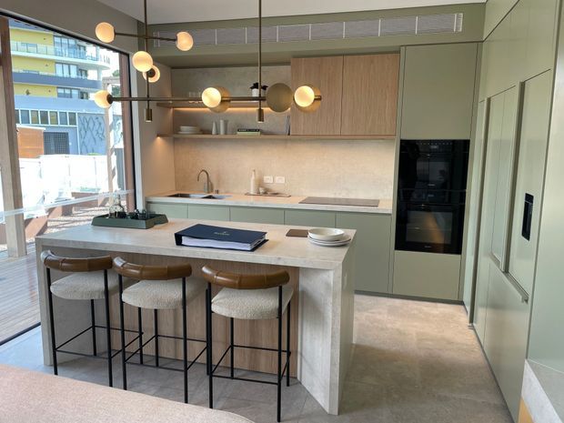 A refined contemporary kitchen with a compact island and three upholstered bar stools. The room features muted sage-green cabinetry, warm timber overhead cabinetry, a pale stone island and benchtop, integrated dark appliances, and a textured neutral splashback. Full-height glazing opens to a balcony or outdoor area, while spherical pendant lights provide warm illumination.