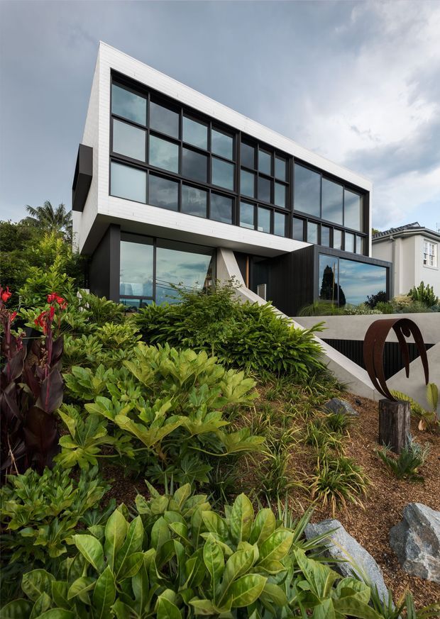 Exterior architectural photograph of a contemporary multi-level home on a landscaped sloping site. The building has a strong rectilinear form, extensive dark-framed floor-to-ceiling glazing, white horizontal cladding, dark wall panels, projecting upper-level volumes, and a sculptural metal garden feature in the foreground. Lush tropical and native-style planting fills the front garden, with a neighbouring traditional house visible at the right.
