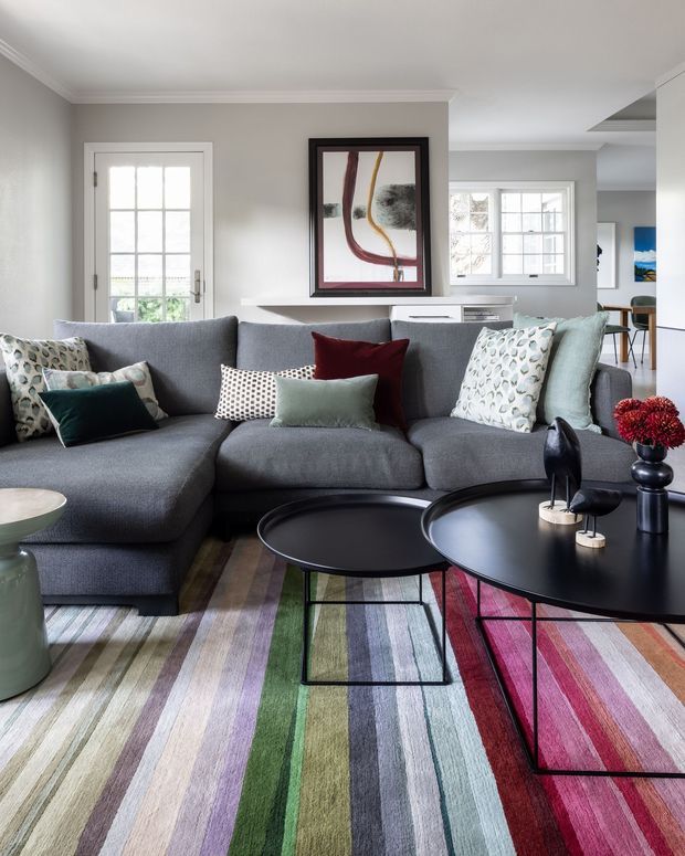 A bright, contemporary living room with a large charcoal-grey sectional sofa, layered patterned cushions, two black oval coffee tables, and a colourful striped area rug. Light grey walls, white trim, French doors, and multiple windows create an airy setting, with abstract framed artwork and a glimpse of a dining area in the background.
