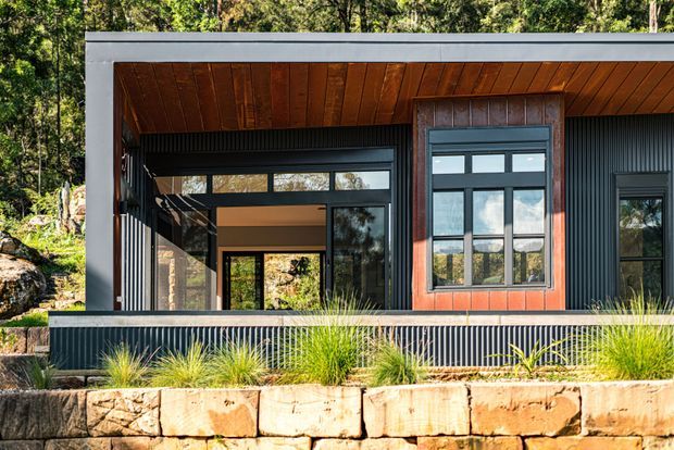Exterior view of a contemporary single-storey building set among trees, featuring dark ribbed metal cladding, a low flat roof, large black-framed glazed openings, and a warm timber-lined soffit. A projecting timber-clad window bay adds contrast, while native-style planting and a rough stone retaining wall form the foreground.