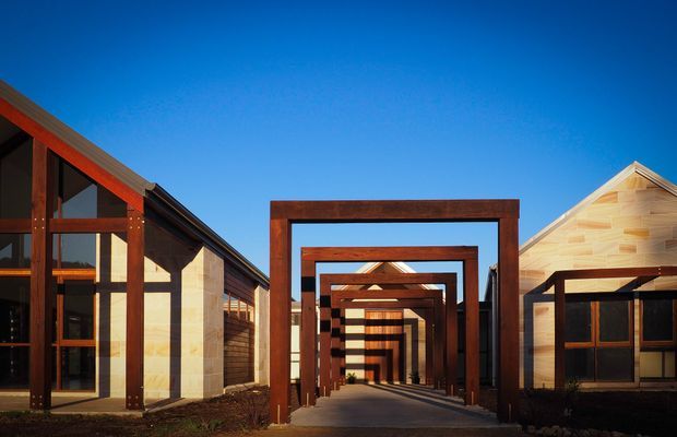 Exterior architectural view of a contemporary timber-clad complex under a clear blue sky. Repeating dark timber portal frames form a covered pedestrian passage between gabled buildings, with extensive glazing and warm-toned wood surfaces creating a strong rhythmic perspective.