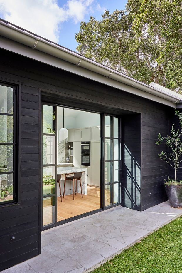 Exterior view of a dark-clad single-storey house with a low pitched roof, gutters, and large black-framed glazed doors opening onto a paved terrace. A bright kitchen with light cabinetry, timber flooring, a marble-look benchtop, and bar seating is visible through the glazing. Mature planting surrounds the building.
