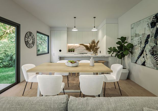 Bright open-plan dining area adjoining a contemporary kitchen and living space. A light timber dining table with six white upholstered chairs sits on pale timber flooring, with white integrated cabinetry, pendant lights, indoor greenery, artwork, and large glazed openings overlooking greenery. The room has a clean, warm, modern feel.