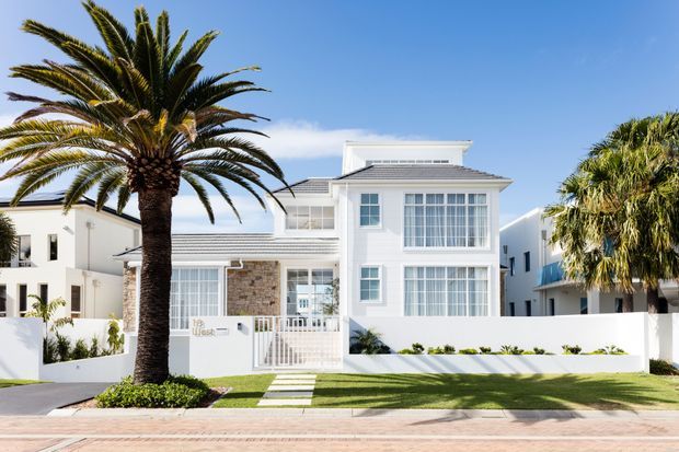Bright front elevation of a contemporary coastal-style two-storey residence with a white rendered facade, grey tiled hipped roof, extensive glazed windows, a small stone-clad feature wall, white boundary walls, and manicured lawn. Mature palm trees and a paved street frontage frame the house.
