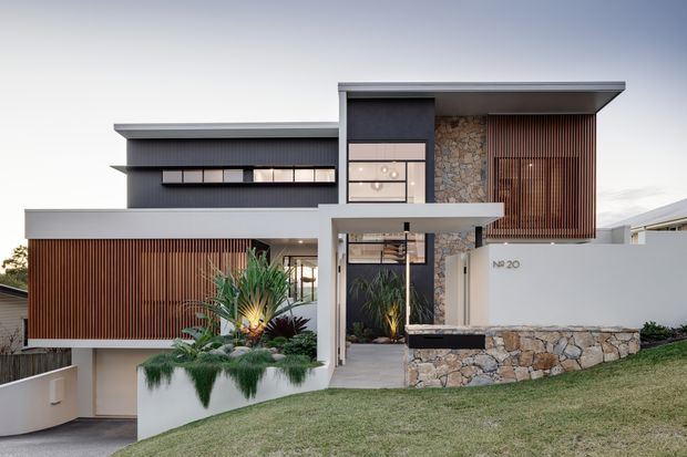 Contemporary multi-level residential facade on a sloping site, featuring dark charcoal cladding, warm vertical timber screening, extensive glazing, pale rendered walls, and prominent natural stone feature walls and retaining walls. The entry is recessed beneath a projecting upper volume, with landscaped raised planters and a manicured lawn in the foreground.