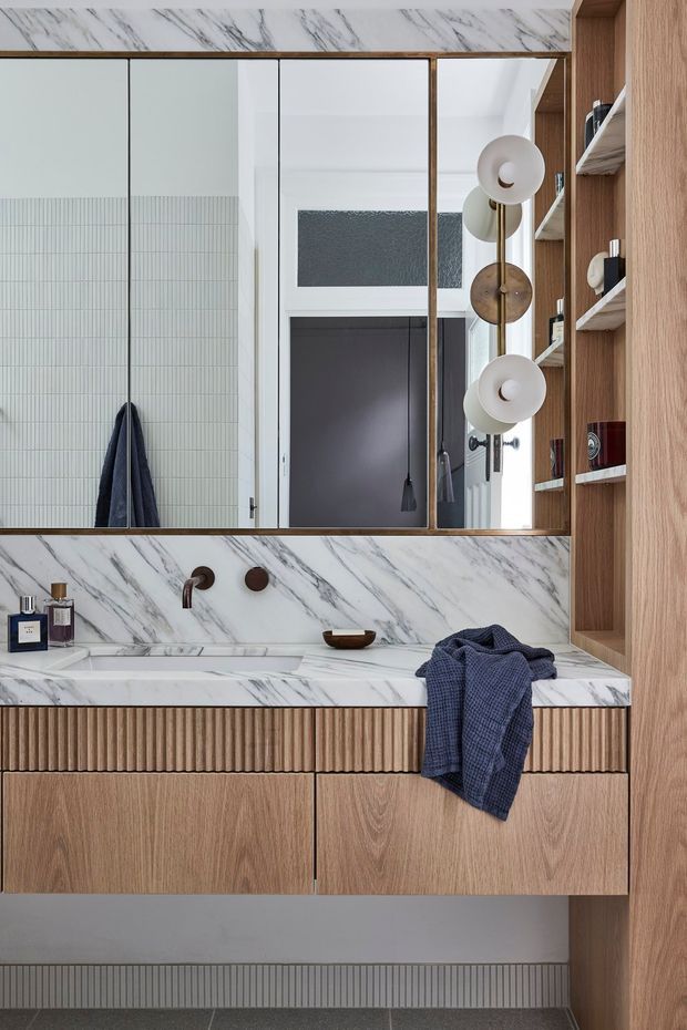 A refined contemporary bathroom vanity with a wall-mounted timber vanity featuring fluted drawer fronts, integrated white marble-look stone benchtop and splashback, brushed bronze wall-mounted tapware, and a large bronze-framed mirror. Vertical open shelving in matching timber occupies the right side, with sculptural globe lighting reflected in the mirror. The room has a restrained palette of white, natural timber, dark grey, and bronze, with a tiled lower wall detail and dark flooring.