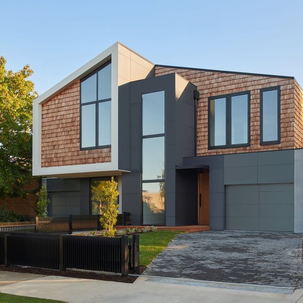 Contemporary two-storey residential facade with a bold asymmetrical composition. The upper level combines warm cedar shingle cladding with dark-framed vertical glazing, while smooth white and charcoal-grey panels define the projecting forms and ground floor. A recessed timber entry, integrated garage, paved driveway, front lawn, and black metal fencing complete the street-facing view.